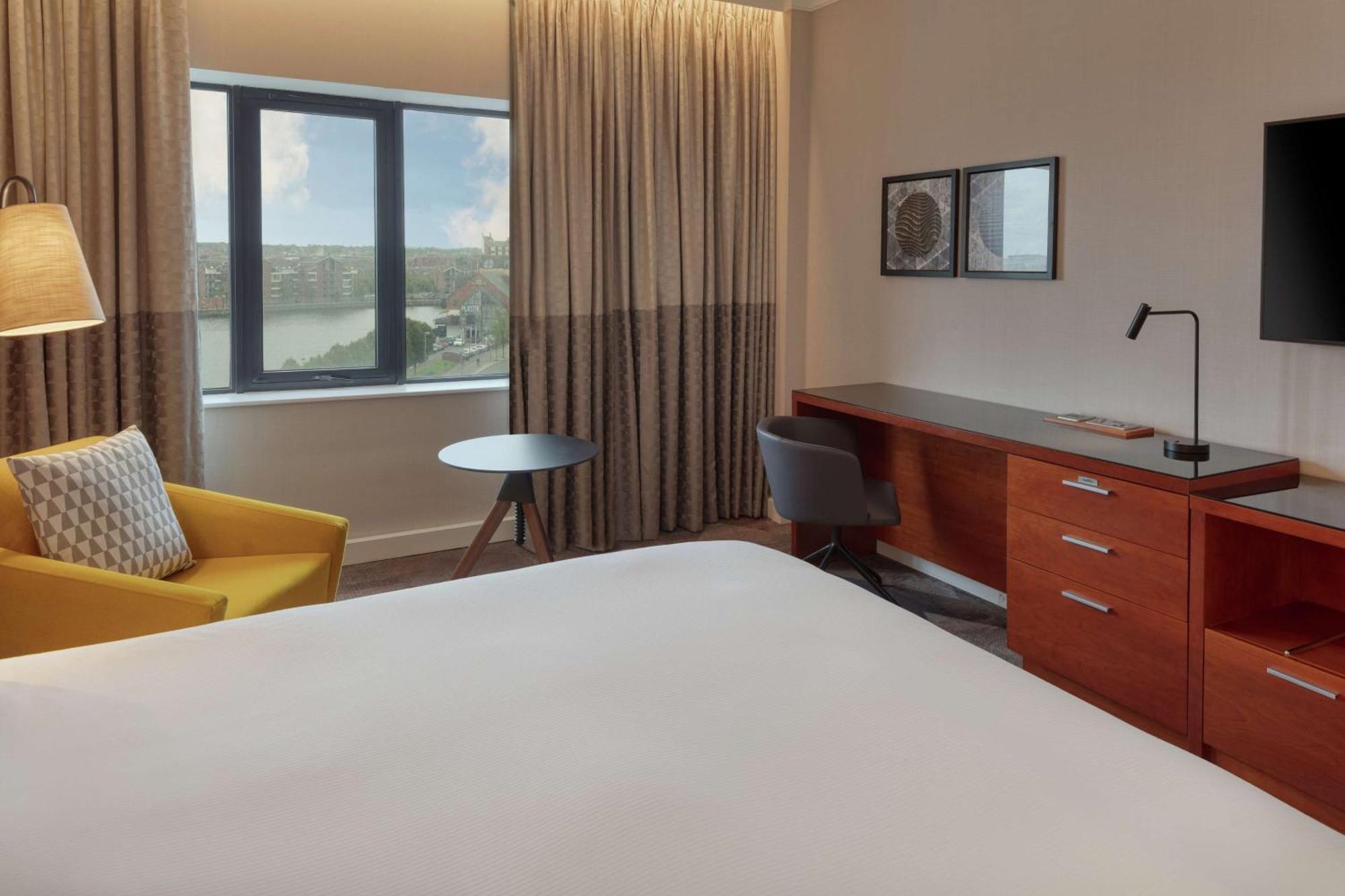 Hilton Belfast Ξενοδοχείο Εξωτερικό φωτογραφία The photo shows a modern hotel room. It features a large bed with a plain white bedspread, and there's a stylish yellow armchair next to it. A small round table is placed nearby, along with a gray chair. The room has large windows covered with light 