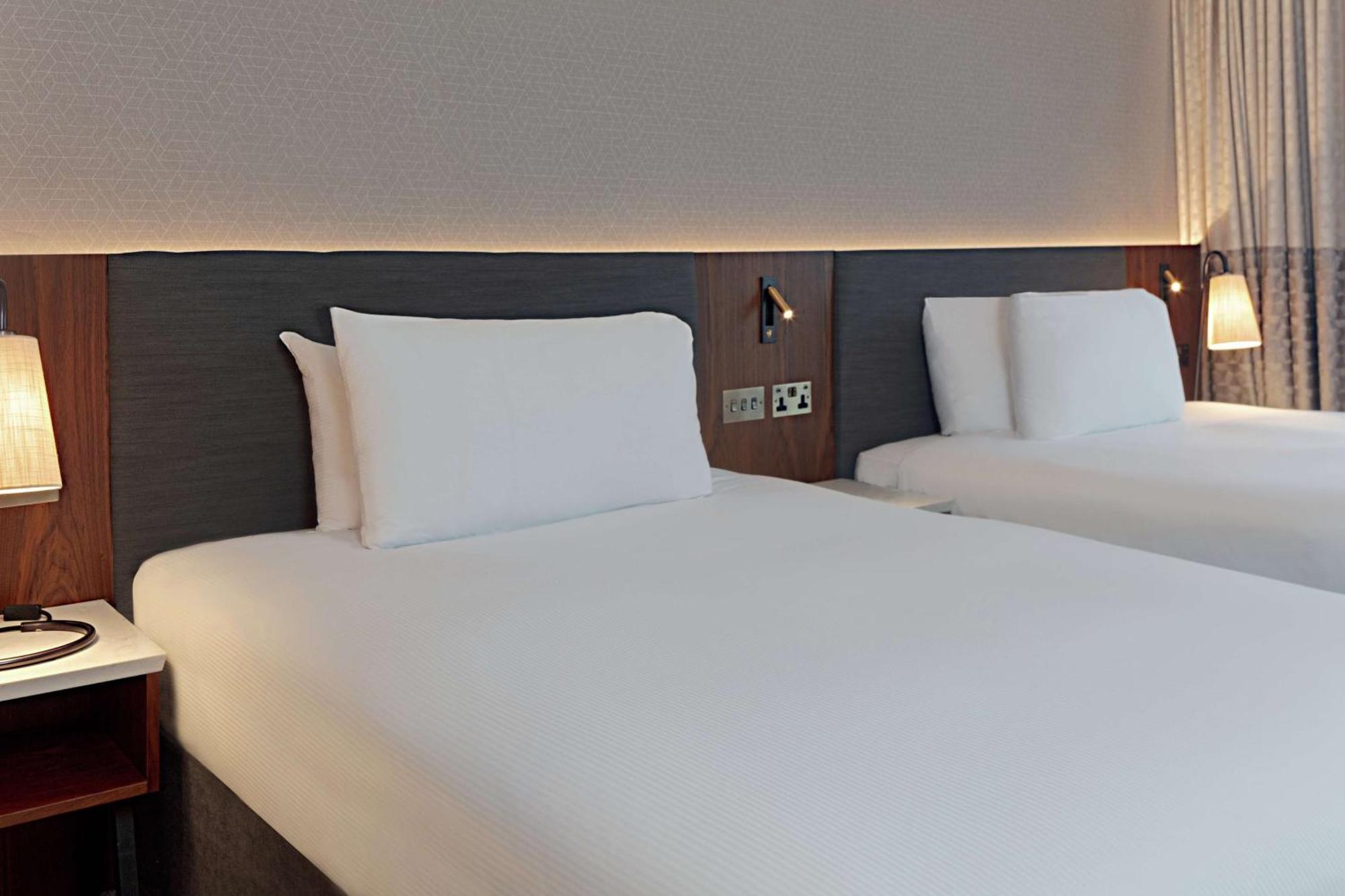 Hilton Belfast Ξενοδοχείο Εξωτερικό φωτογραφία The photo shows a modern hotel room with two neatly made beds. Each bed features white linens and pillows, with a simple yet stylish headboard in a dark wood finish. The room has a soft, ambient light illuminating the area, creating a welcoming atmos