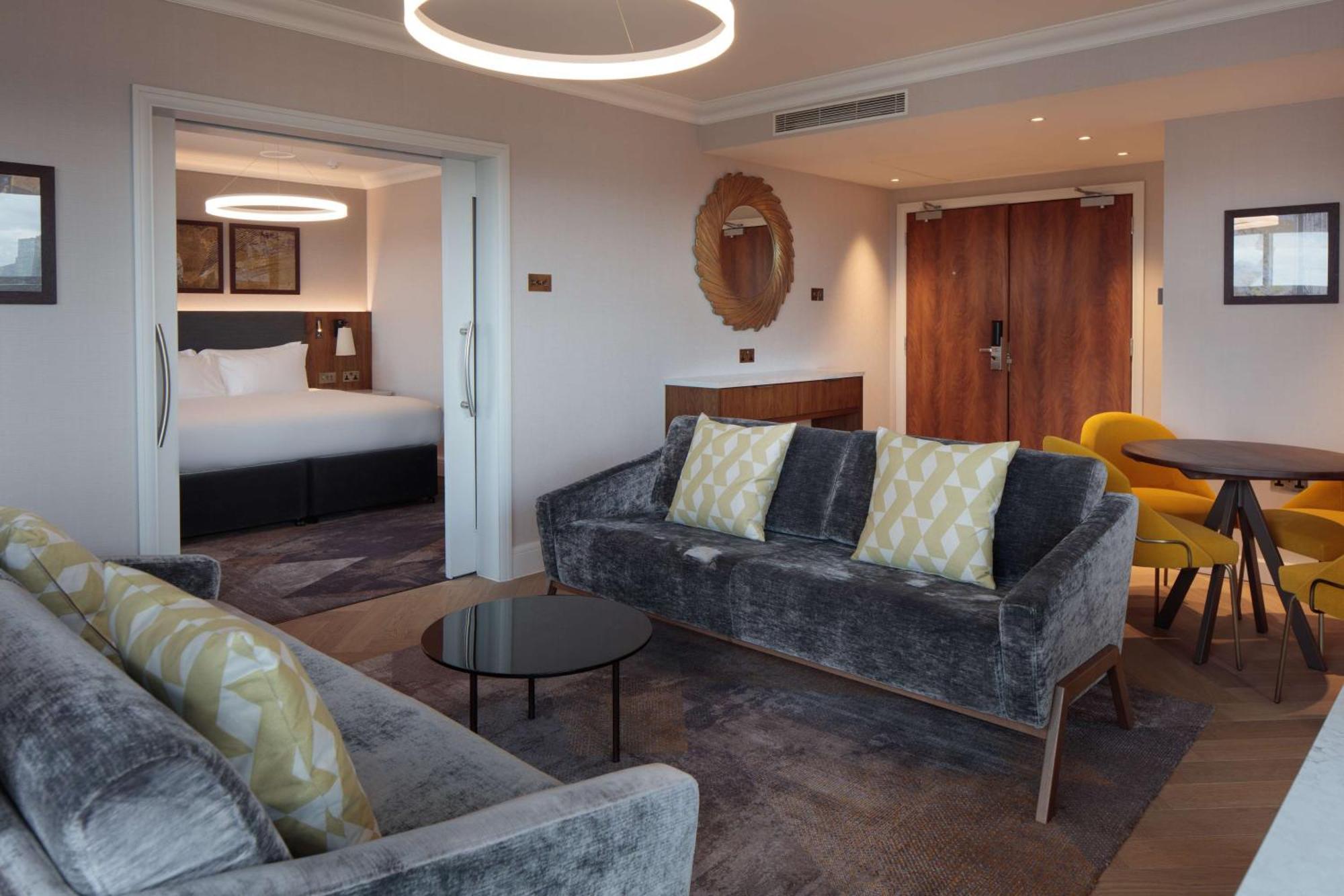 Hilton Belfast Ξενοδοχείο Εξωτερικό φωτογραφία The photo shows a modern and stylish hotel suite. In the foreground, there is a comfortable gray sofa with decorative pillows and a small black coffee table. Adjacent to the sofa is a dining area with a round table and yellow chairs. In the backgroun