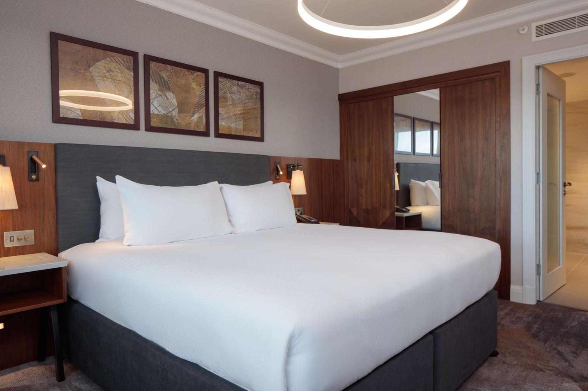 Hilton Belfast Ξενοδοχείο Εξωτερικό φωτογραφία The photo shows a modern hotel room with a clean and contemporary design. The focal point is a large bed with crisp white bedding and two fluffy pillows. The headboard is gray, and next to the bed, there are stylish bedside lamps. Above the bed, ther