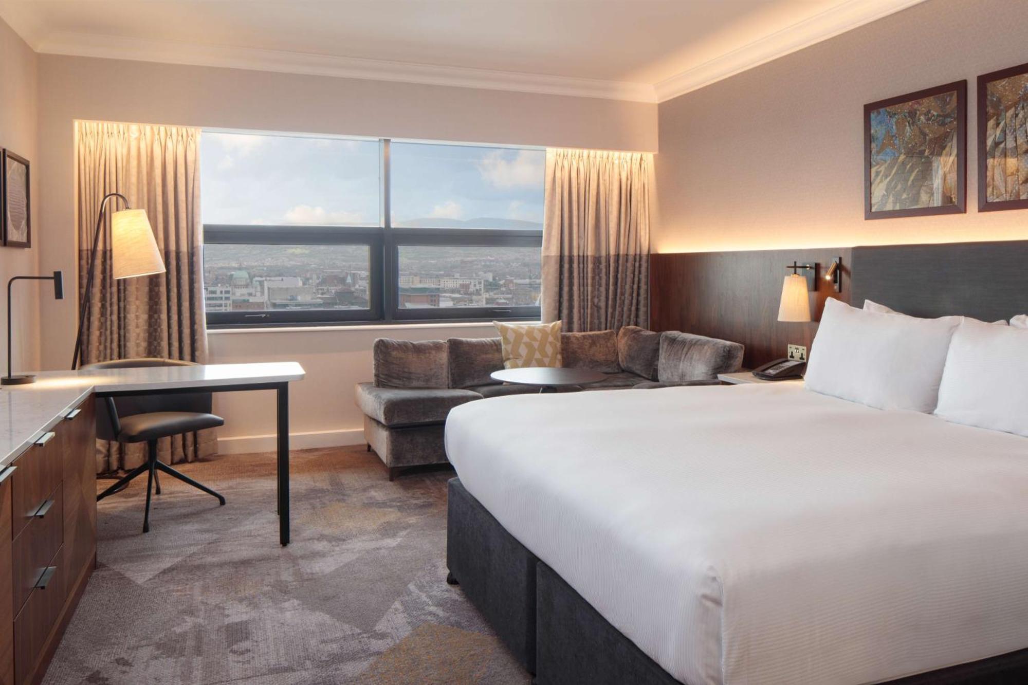 Hilton Belfast Ξενοδοχείο Εξωτερικό φωτογραφία The photo shows a modern hotel room featuring a large bed with white linens positioned against a wall. On one side, there is a small seating area with a couch and a round table. A desk and chair are also present, suggesting a workspace. The room has 