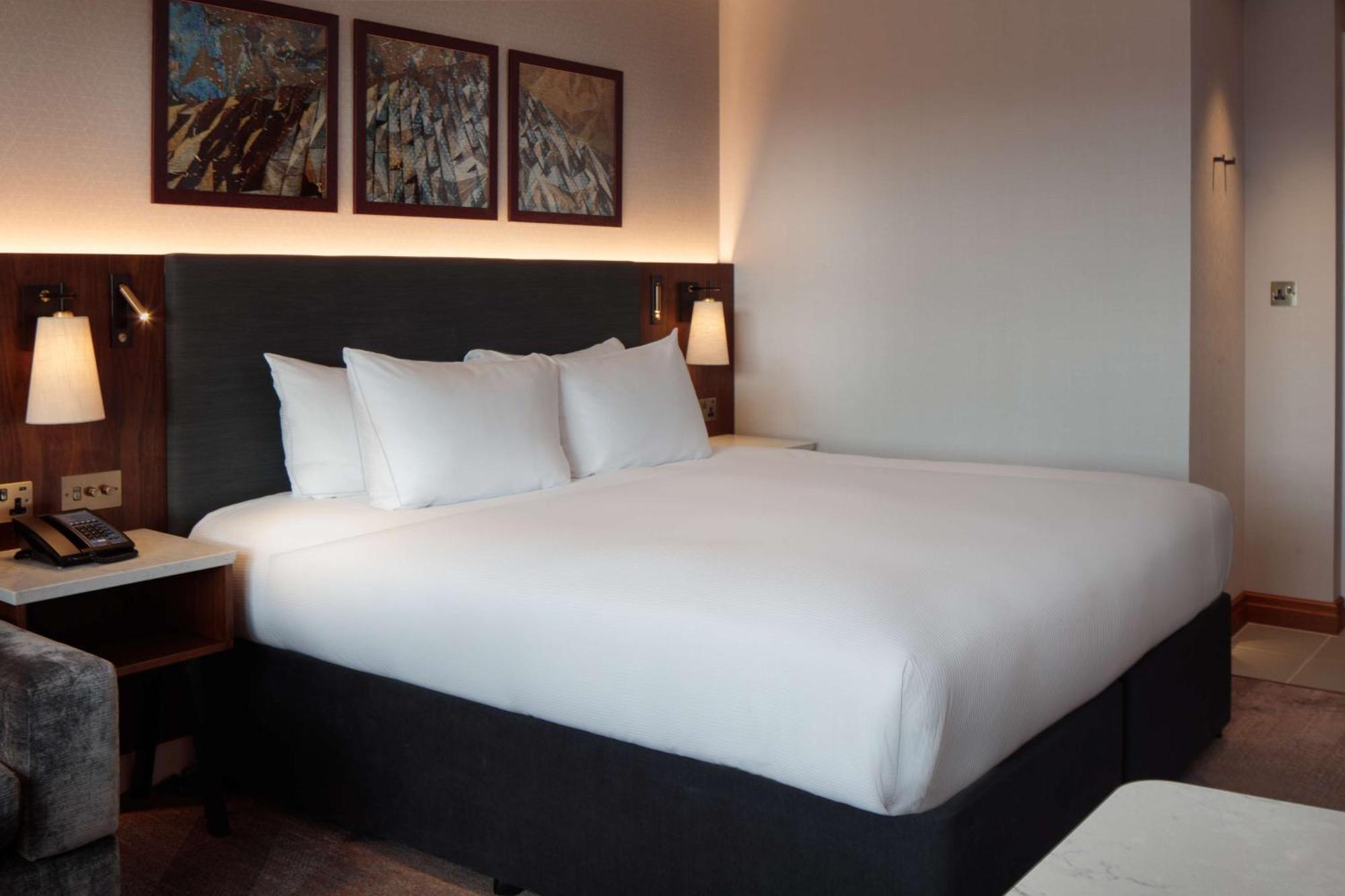 Hilton Belfast Ξενοδοχείο Εξωτερικό φωτογραφία The photo shows a modern hotel room featuring a large bed with white bedding and several pillows. The bed is positioned against a dark-colored headboard. On the wall above the bed, there are three framed artworks. The room has soft lighting, and ther