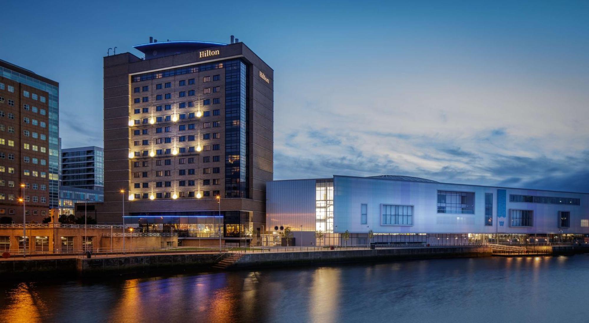 Hilton Belfast Ξενοδοχείο Εξωτερικό φωτογραφία The image shows a Hilton hotel building along a waterway, illuminated during twilight. The hotel features a modern architectural design with multiple stories and large windows. To the right of the hotel, there is another structure that appears to be 